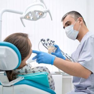 Dental professional in mask and blue gloves shows a dental model to seated patient in modern clinic with overhead light