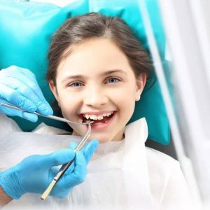 Young girl in teal dental chair smiling while dentist examines her teeth with instruments in professional clinic setting